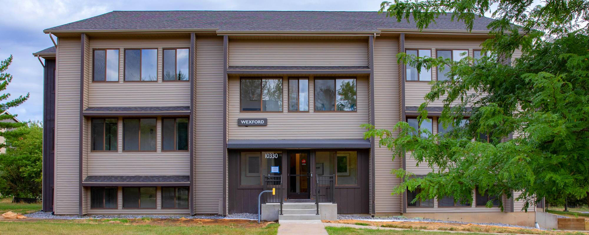Three-story Grand Valley apartment building with brown siding and large windows, surrounded by greenery.